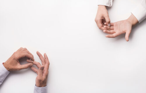 Two pairs of hands on a white background, each person removing a wedding ring from their finger, symbolizing separation or the waiting period divorce often requires.