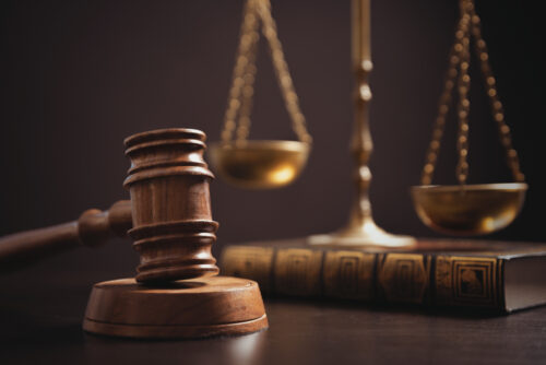 A wooden judge’s gavel rests on a sound block in front of a law book, with golden scales of justice blurred in the background, symbolizing law and legal proceedings.