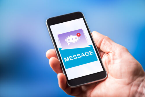 A hand holding a smartphone displaying a screen with a chat bubble icon, a notification alert, and the word "MESSAGE" in large letters. The background is blurred blue.