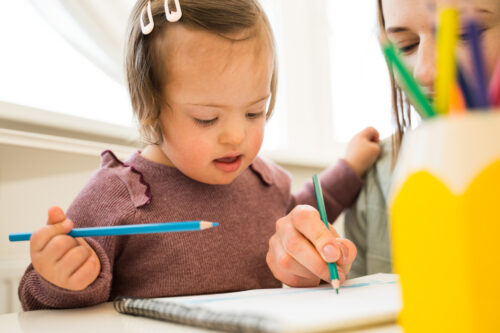 A young girl with Down syndrome draws with colored pencils in a notebook, sitting beside an adult who is helping her. A yellow pencil holder with pencils is visible in the foreground.