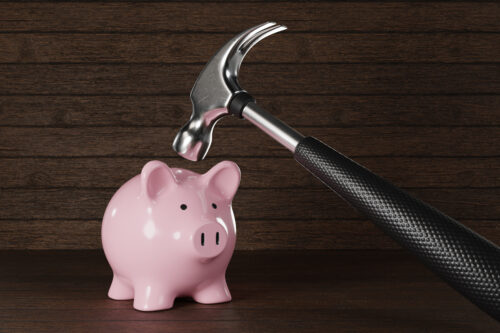 A pink piggy bank sits on a wooden surface while a hammer is raised above it, as if about to strike. The background consists of dark wooden planks.