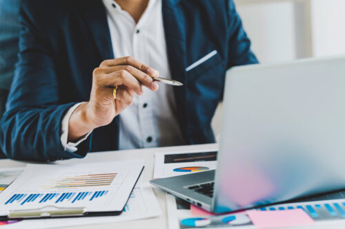 A person in a suit holds a pen while working at a desk with a laptop, charts, and graphs, suggesting a business or financial analysis setting.