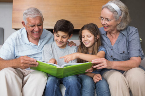 Two older adults and two children sit together on a couch, smiling and looking at a large green book, as one child points at a page while the others watch with interest.