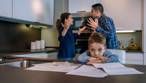A young boy looks sad at a kitchen table covered with papers, while two adults argue in the background, both raising their hands in frustration. The scene suggests family stress or conflict over finances.