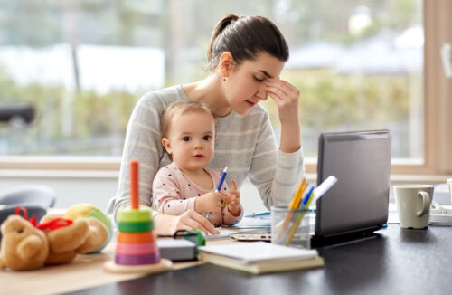 A tired woman sits at a table with a laptop, holding her head and paperwork, while a baby sits on her lap holding a pen. Toys are scattered on the table, highlighting the challenges of balancing work, childcare, and supervised visitation.