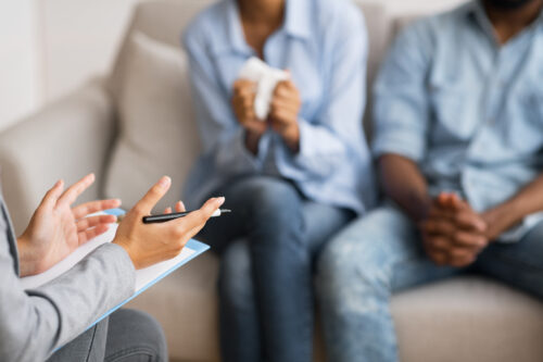 A counselor gestures with a pen while talking to a couple sitting on a couch. One person holds tissues, and both appear to be listening intently, suggesting a private divorce counseling session.